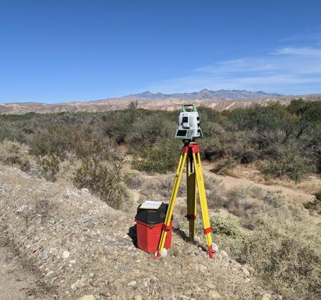 Terrestrial laser scanner on a yellow tripod in a floodplain with brush, distant mountains, and blue sky. 