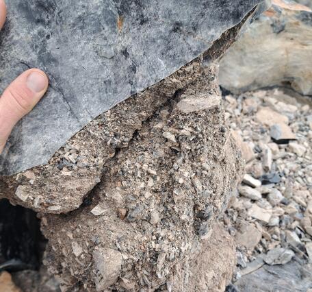 A photograph of a fracture filled with light grey ash and multi-colored rock fragments that is cutting through dark obsidian rock. A finger included for scale indicates that the fracture is about the width of a human hand, and some of the largest angular rock fragments are finger-sized. In the background, a rubble-covered surface of the Panum lava dome is visible.