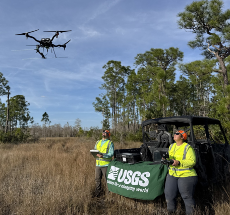 two people in yellow vests lean against an OHV, look up at drone flying above them, grasses and trees in background