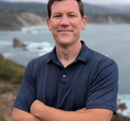 A man in a blue t-shirt with short brown hair stands along a coastline.