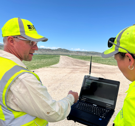 two people in bright yellow USGS gear chat outside while looking at a small computer screen
