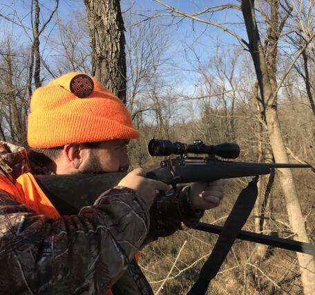  hunter wearing a blaze orange beanie and camouflage jacket aims a bolt-action rifle with a scope.