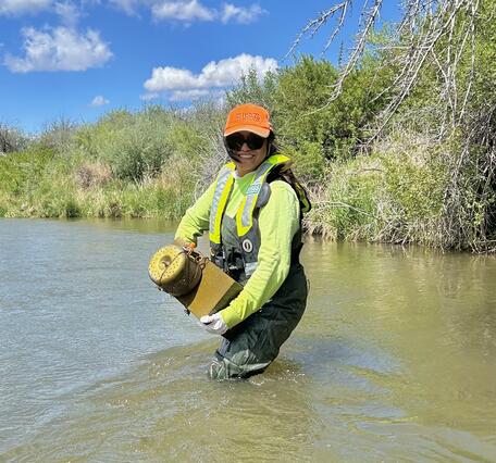 A person wearing waders, PFD, and a USGS hat is standing knee-deep in a river holding a passive pesticides sampler.