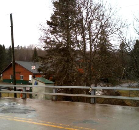 USGS streamgage housing behind bridge guardrails, with a wet road surface on a rural bridge in the foreground.