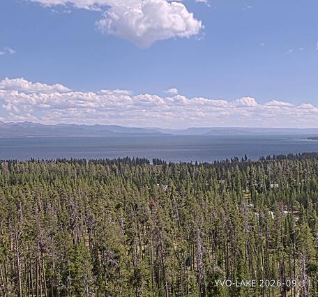 forested area in teh foreground, lake in the middle around, and mountains in the background under partly cloudy sky