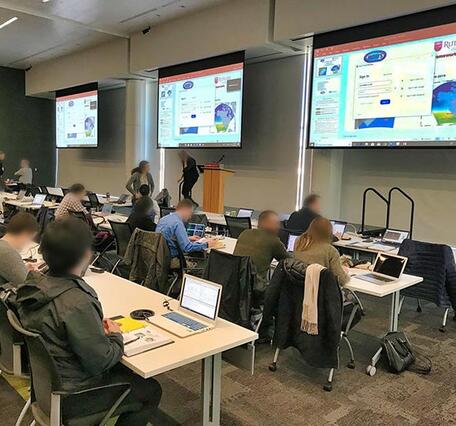 Training room with tables and large screens. Participants sit at the tables.