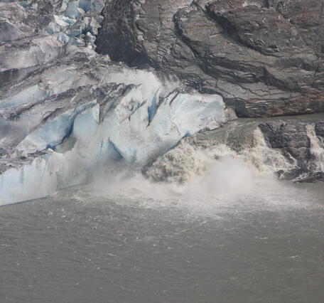 White, frothy water pours from the bottom of a blue glacier, marring the surface of a calm grey lake.
