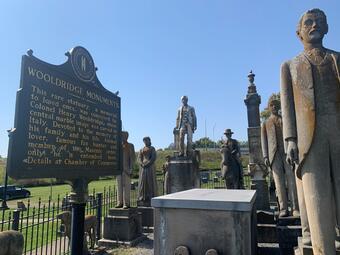 Historical marker installed beside weathered human-figure statues, all enclosed by an iron fence.