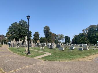 A set of weathered statues visible from a cemetery road intersection, with a pathway leading up to the burial plot.