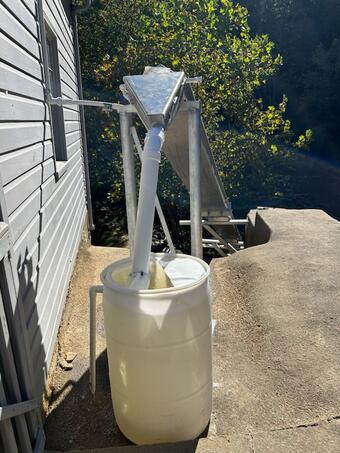 A white barrel sits beside a building, with PVC pipes feeding into it from a metal chute above.