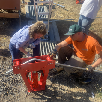 two people crouch near a pipe bender while feeding 1 inch metal pipe through