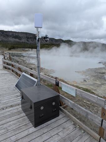 Webcam on a mast attached to a battery box on a boardwalk overlooking a murky and steaming pool