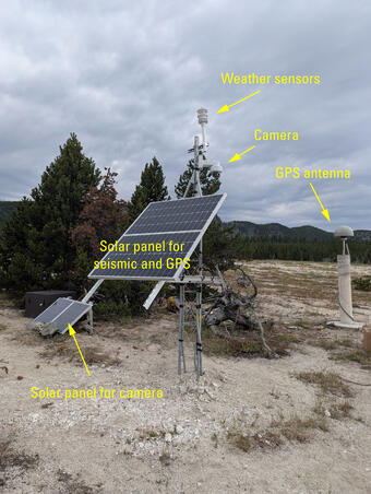 barren ground with a few small pine trees and a metal tower with a solar panel and other sensors