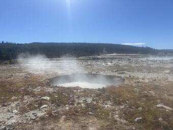 Circular pool of boiling water in a barren area strewn with rocks with a forested ridge in the distance under blue sky.
