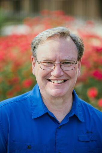A man in a blue collared shirt wearing glasses and seated in front of red flowers.