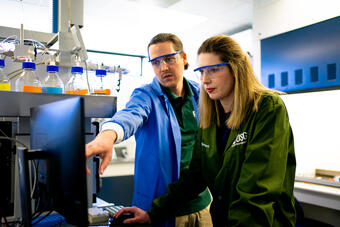 In a lab, two scientists wear lab coats and glasses. One scientist uses a computer as the other one points to the screen.