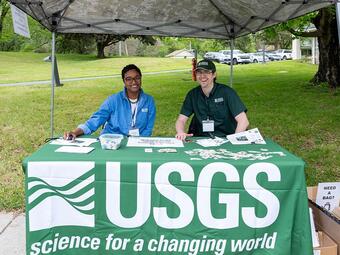 Two USGS employees smile while sitting behind a USGS tablecloth covered table under a pop up tent