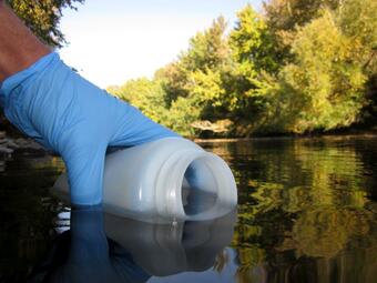 a person wearing an exam glove holding a plastic bottle partially submerged in a river