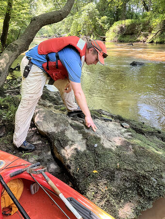 A usgs geologist in a bright red PFD examines a large boulder along a creek. 