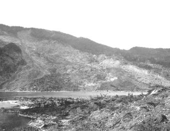 Black and white photo of a hillside that has been devastated by a landslide, with rock debris in the foreground and a lake