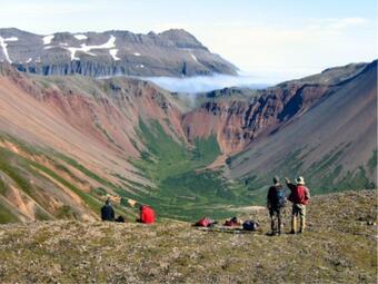 Two people standing by a ledge facing and gesturing away towards mountains in the distance; packs and two people on ground