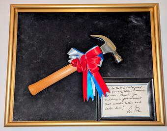 Hammer with a red, white, and blue bow mounted on black felt canvas with a golden frame