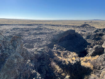 fissure in dark rock with dry grass, and dry grassy landscape in the distance under blue sky
