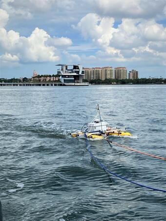 A collection of equipment is mounted on yellow catamaran in the bay. In the background: a pier & skyline with tall buildings