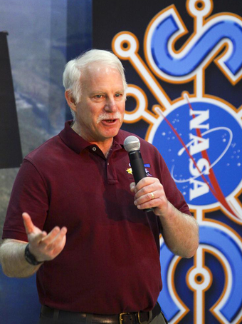 A man speaking into a microphone with some colorful banners behind him.