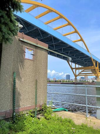 Streamgage housing next to a river with a large suspension bridge in the background