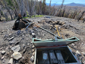 In this photo of a bare, rocky hill in the middle of a fire-blackened forest, a pole and strapping are laying on the ground next to a charred stump. In the foreground, a dented and warped box contains the melted remains of several large bits of electronics. USGS photo by Ryan Presser, NCSN.