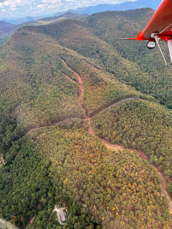 An aerial photo of green mountains with a landslide cutting through the trees. A red plane wing can also be seen. 