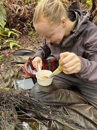 Person sitting on ground in rain gear holding a sample in a plastic cup and a baster.