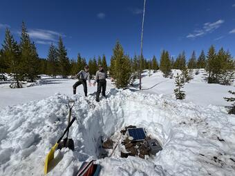 Two woman point out a pit in snowy landscape with equipment in the pit. Sparse trees and blue sky in background.