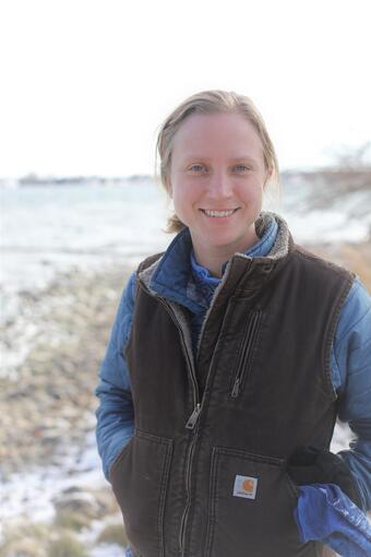 A woman with blonde hair in a long sleeved shirt and vest stands for her picture. 