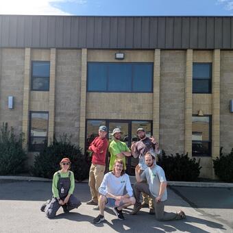 Six people pose in front of a brown brick building on a sunny summer day