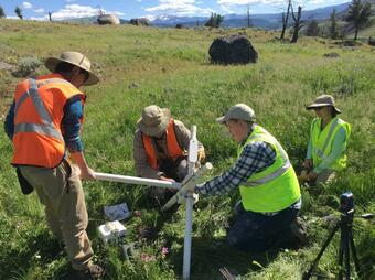 Four scientists in safety vests in a grassy meadow with occasional bounders setting up geophysical equipment