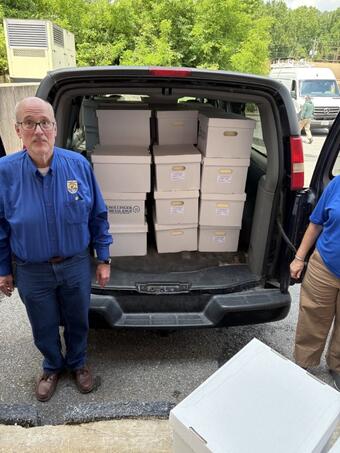 Two workers in blue shirts stand next to the open back doors of a van filled with cardboard record boxes. 