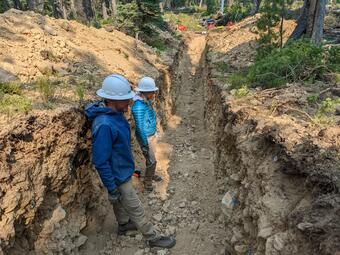 Two people standing in a shallow trench a few feet wide and deep in a forested landscape. They are examining the trench wall