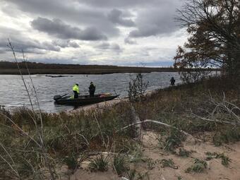 Two people standing in a boat near a sandy shoreline of a slough when other person stands on a the shore