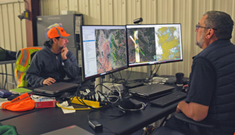 Two people in a hangar at a workstation displaying hyperspectral survey maps, surrounded by laptops and field equipment
