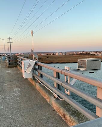 Looking along a bridge rail where a radar unit is suspended out over the river and tied into a control box with a rain gage