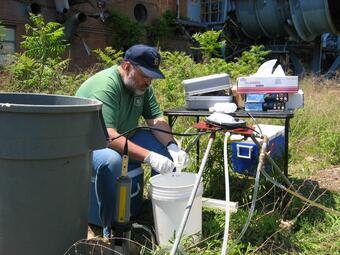 USGS scientist sitting on a chair in a field collecting water from a well into a bucket