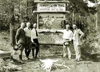Five men in 1920s-era suits stand in front of a wooden sign for Howard Eaton Trail; forest behind and bison skull in front