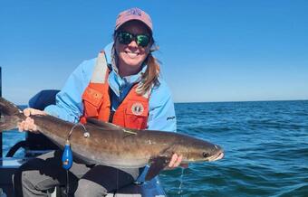 A peson in a hat and sunglasses and orange life jacket leans on the side of a boat holding a cobia