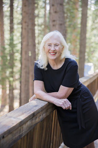 Kass Green leans against a wood railing, smiling at the camera, with a forest in the background