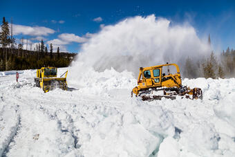 Snowplow removing snow form a road, with trees in the background under partly cloudy skies