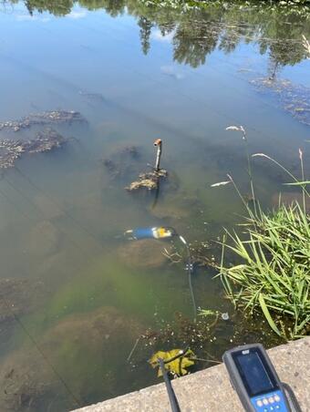 A blue EXO sonde submerged in shallow muddy lake water connects by a cable to the handheld device that shows the readings