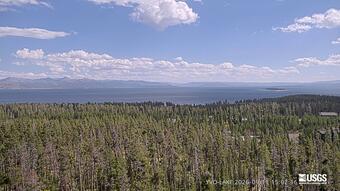 forested area in teh foreground, lake in the middle around, and mountains in the background under partly cloudy sky