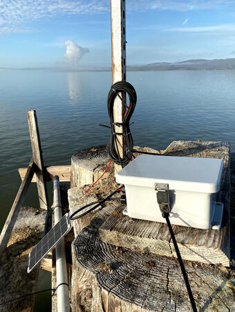 A nondescript white instrument box sits up high atop piers in the water with cables and a small solar panel.